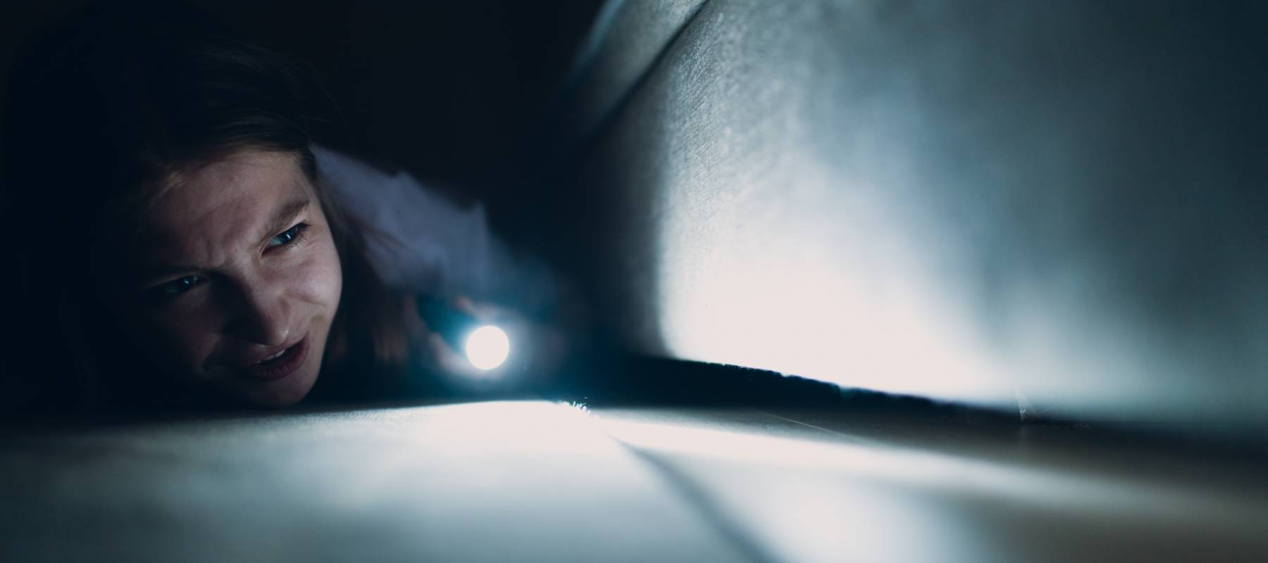 A woman shining a flashlight under the sofa.