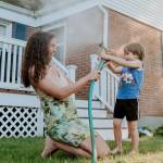 Woman with curly hair and young boy play with a hose outside an older home