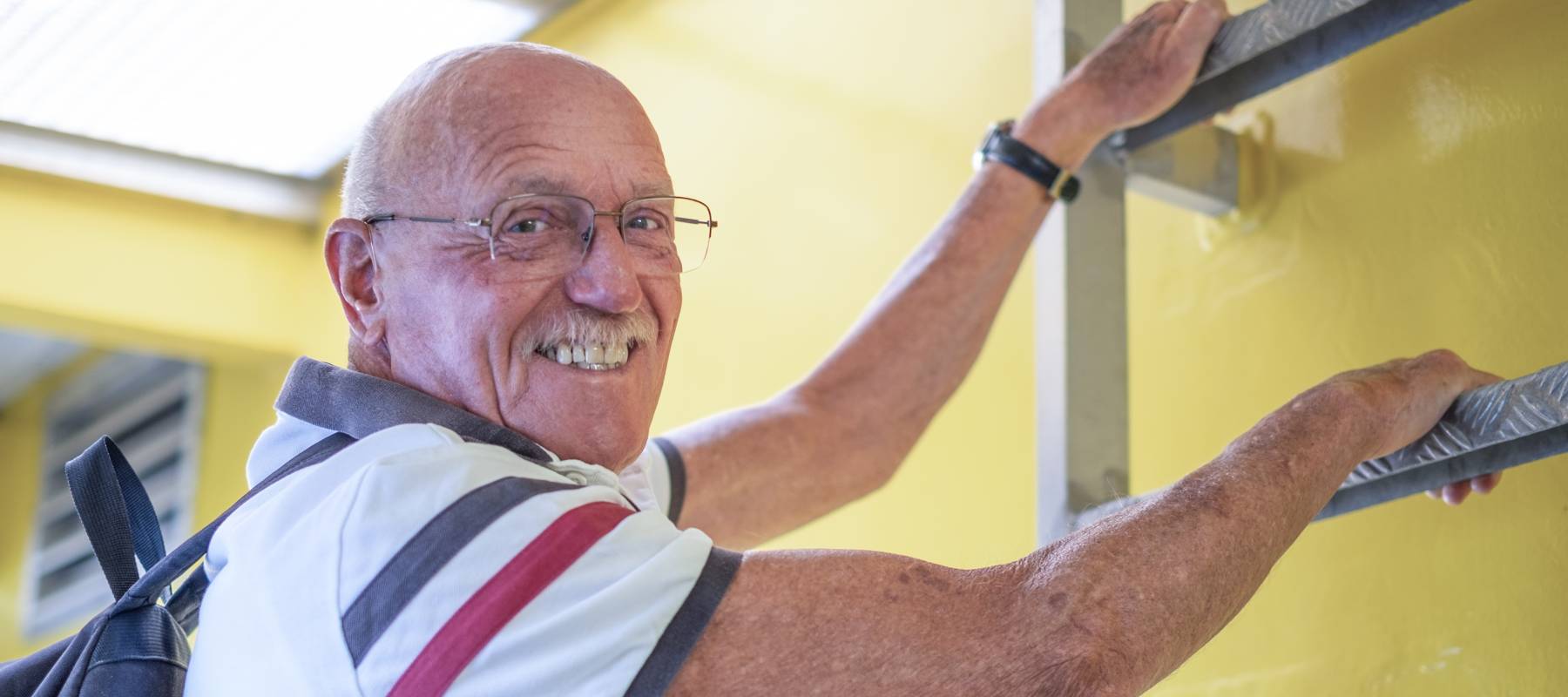 smiling Caucasian senior man with backpack on shoulders preparing to climb an emergency ladder