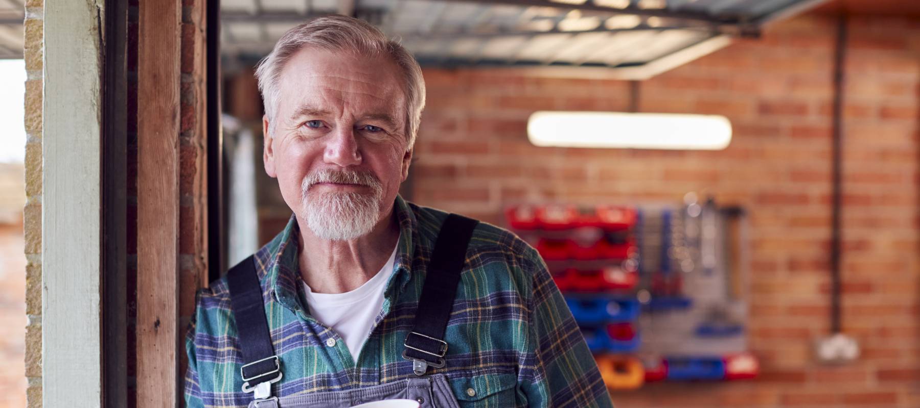 senior Caucasian man in overalls smiling at the camera with a cup of coffee in a mug