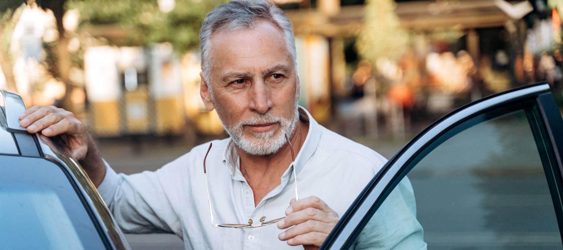 senior Caucasian man is standing near his rental SUV car and holding sunglasses