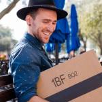 Hipster wearing a wide-brimmed hat sitting on a park bench holding a box from Amazon