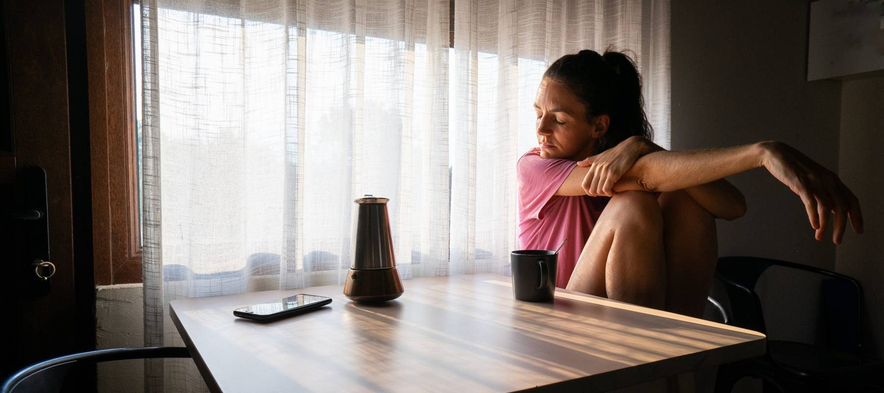 Sad woman sitting at table with smartphone and coffee pot by window