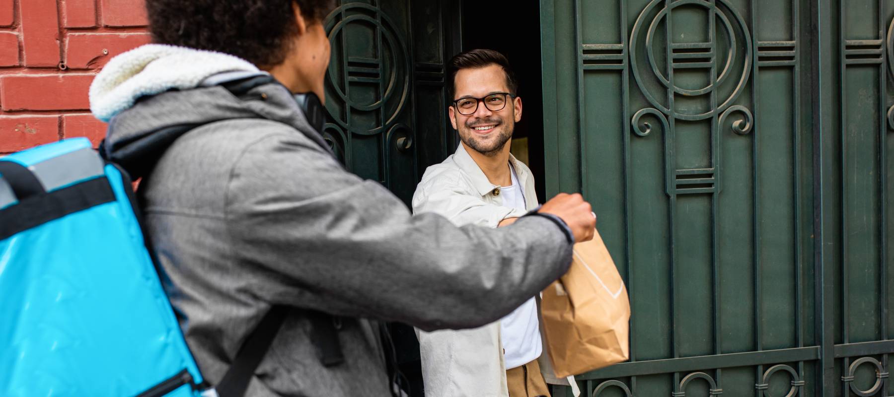 Male courier with bicycle delivering food in brown paper bag to the customer at a doorstep in a city.