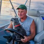 Man driving pontoon boat with dog in his lap.