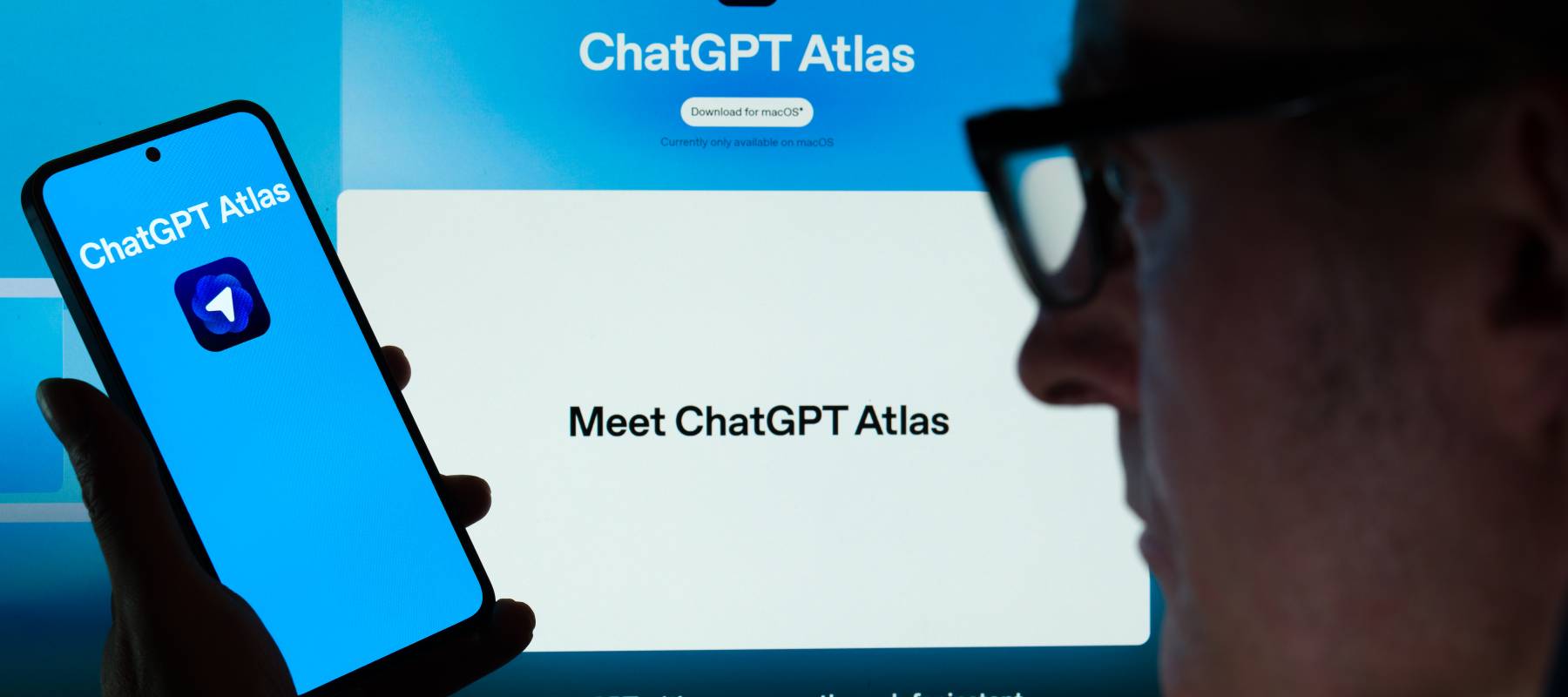 ChatGPT Atlas icon is displayed on a mobile phone held by a man with a screen in the background also showing the ChatGPT Atlas logo.