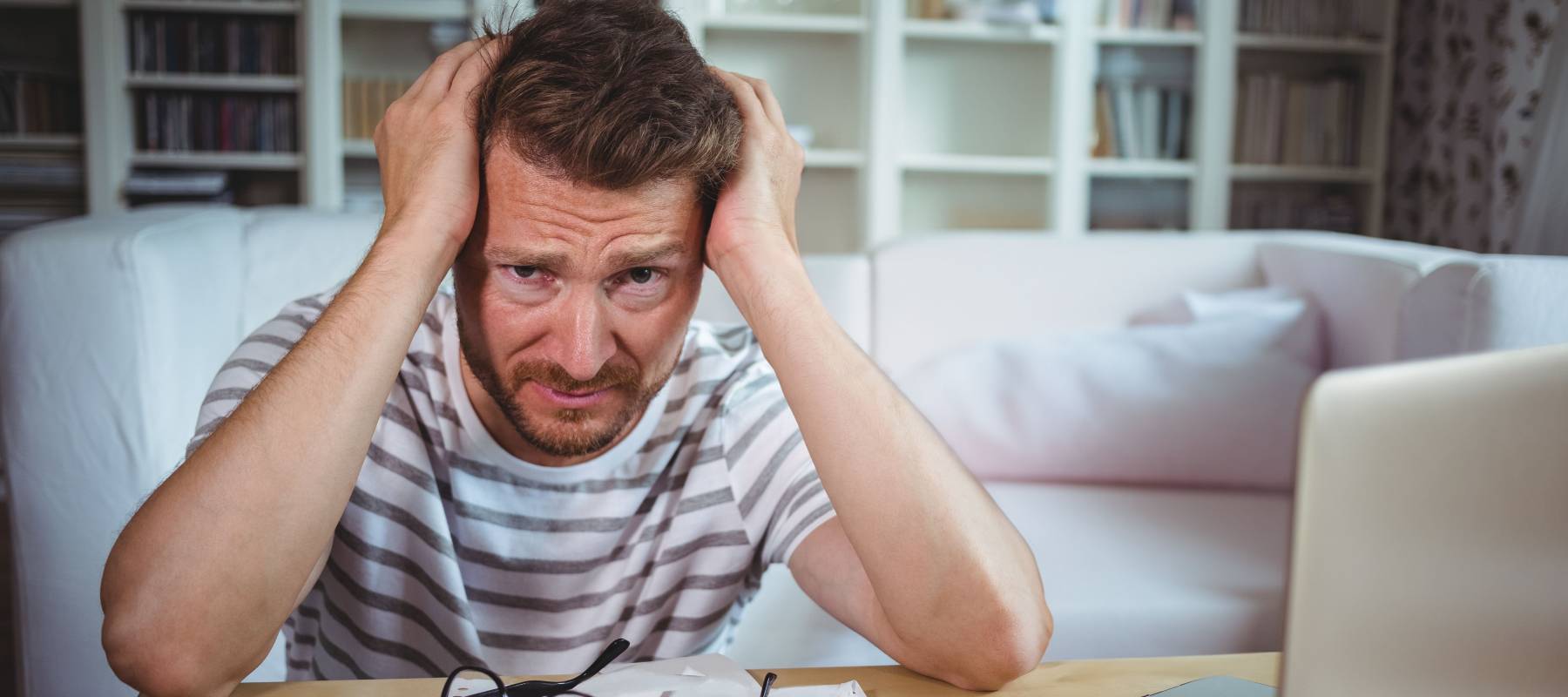 Mid-adult man leaning at table at home holding head amid papers, glasses, calculator, and laptop.