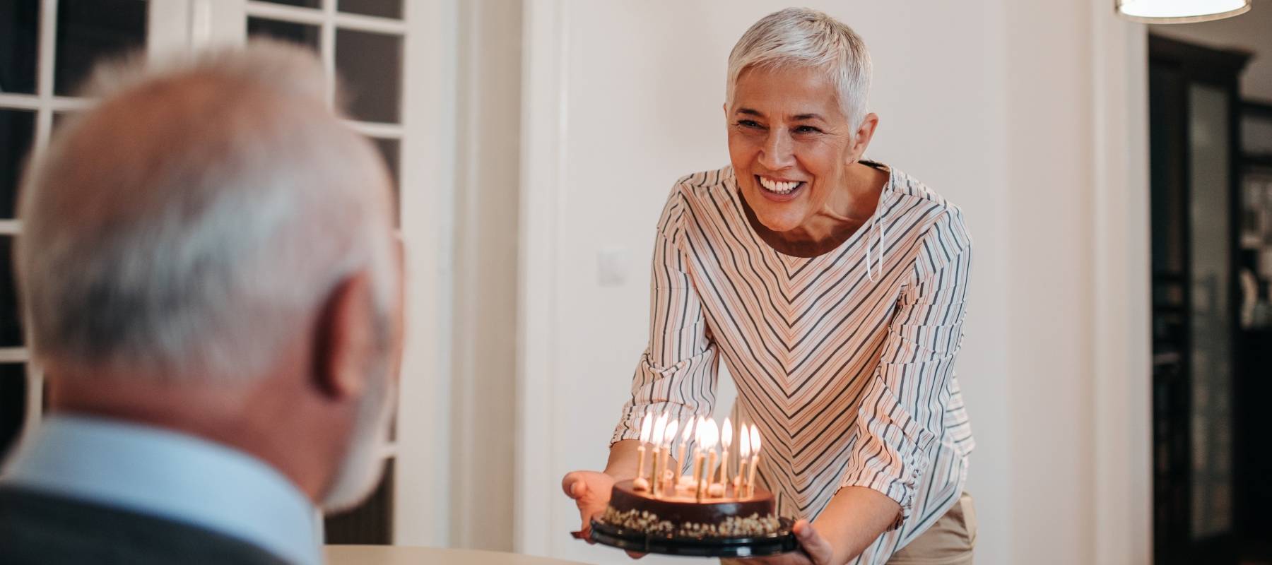 Smilling woman approaches her husband with a birthday cake lit with candles.
