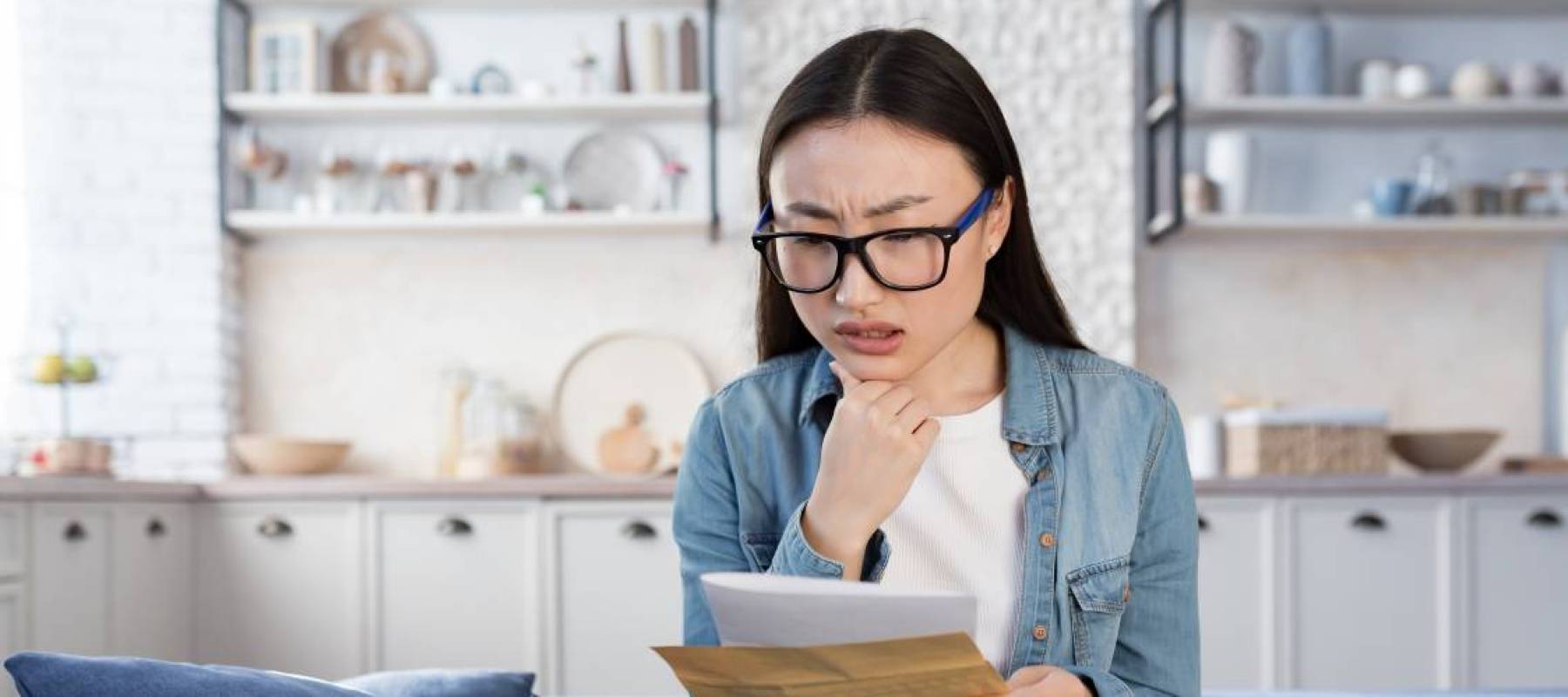 young Asian woman holding paperwork on her couch, looking concerned