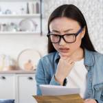 young Asian woman holding paperwork on her couch, looking concerned