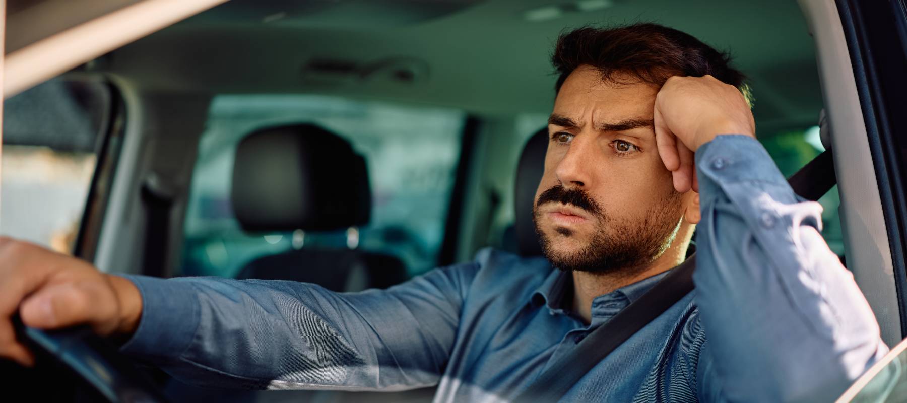 A frustrated man stuck in traffic looks askew at the camera with one hand resting on the wheel.