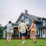 Real Estate Agent Showing a Beautiful Big House to a Young Successful Couple. People Standing Outside on a Warm Day on a Lawn, Talking with Businesswoman, Discussing Buying a New Home.