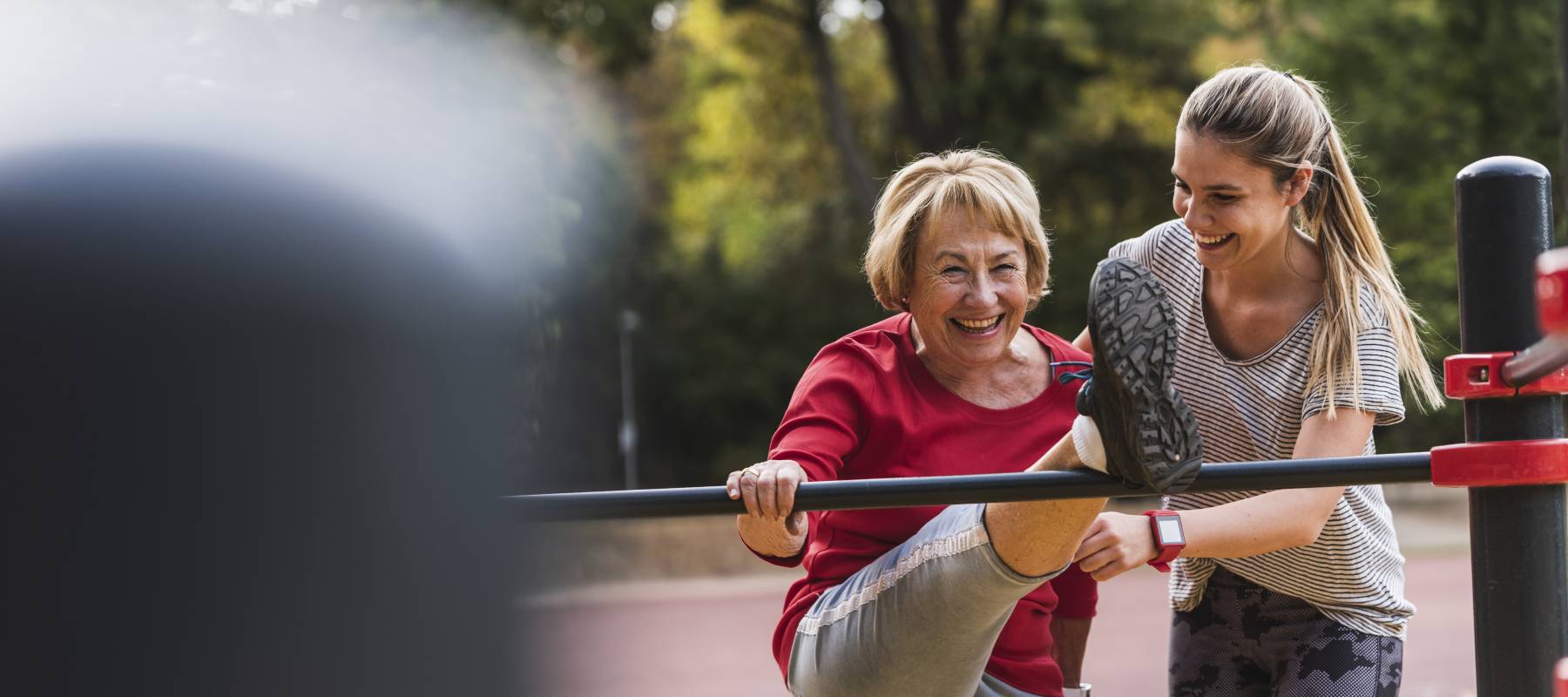 A 20-something daughter helping her mid-60s mother stretch on a playground.