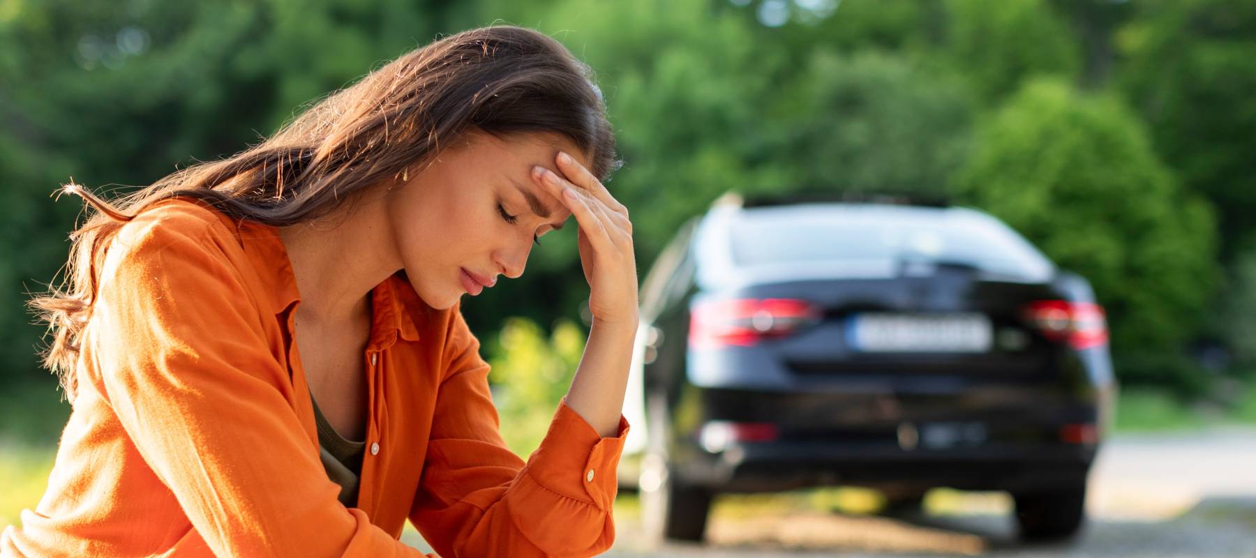 A young woman with medium-length brown hair and a bright orange blouse crouches at the side of the road with her head in her hands, and her car in the background.