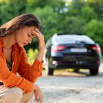 A young woman with medium-length brown hair and a bright orange blouse crouches at the side of the road with her head in her hands, and her car in the background.