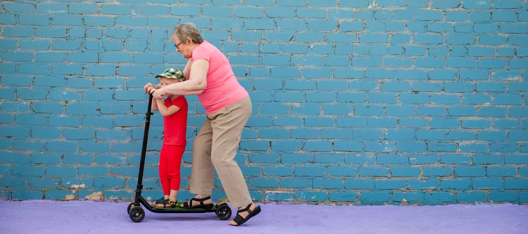 Elderly woman in pink T-shirt and glasses with her little grandson riding scooter.