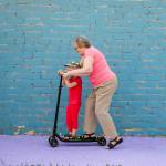 Elderly woman in pink T-shirt and glasses with her little grandson riding scooter.