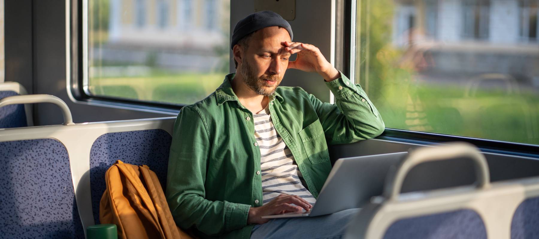 Weary man works on laptop on train with his hand on his head to shield from the sun.