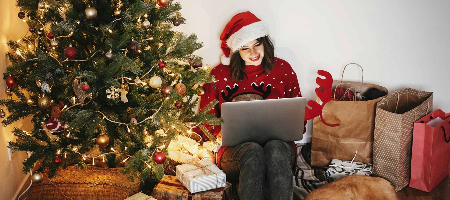 happy girl in santa hat shopping online on laptop and sitting with cute dog at golden beautiful christmas tree with lights and presents in festive room. christmas sale and discount concept