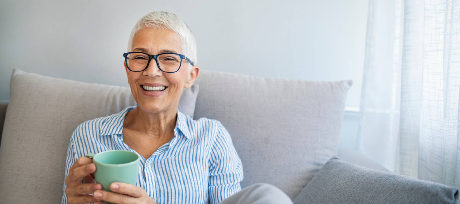 a Caucasian senior woman smiling at home on her couch, holding coffee