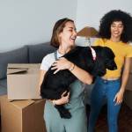 Moving, lesbian and couple with dog in new home, boxes and smile in real estate together. P