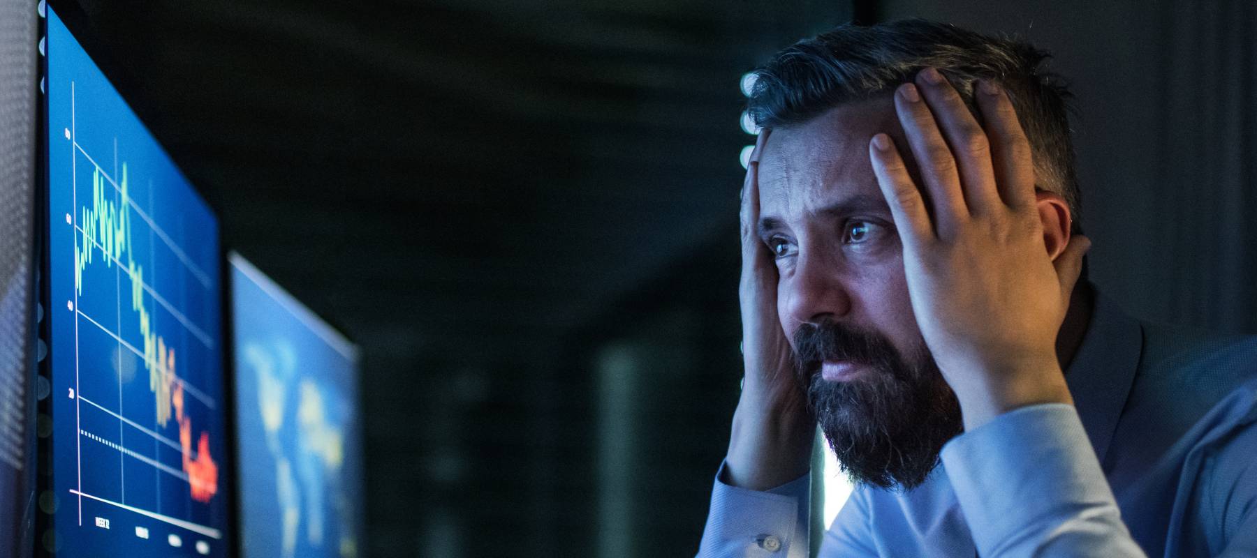 A bearded man in a dress shirt, lit only by the glow of his monitor stares, at flatlining stocks on a triple-monitor set up.