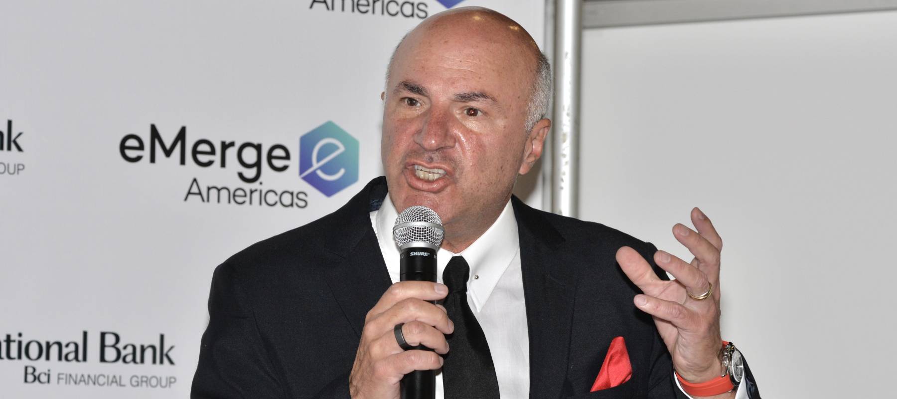 Kevin O'Leary speaks into a microphone in a black suit with a red pocket square.