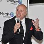 Kevin O'Leary speaks into a microphone in a black suit with a red pocket square.