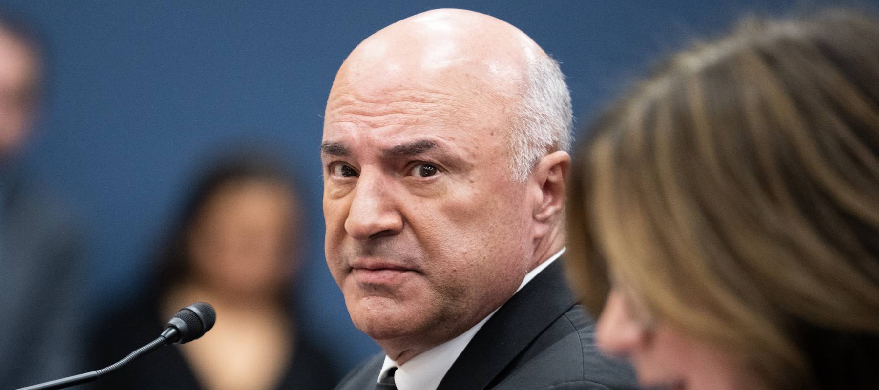 Kevin O'Leary looks askew at the camera in a black suit, black tie and white shirt with a deep expression of concern.