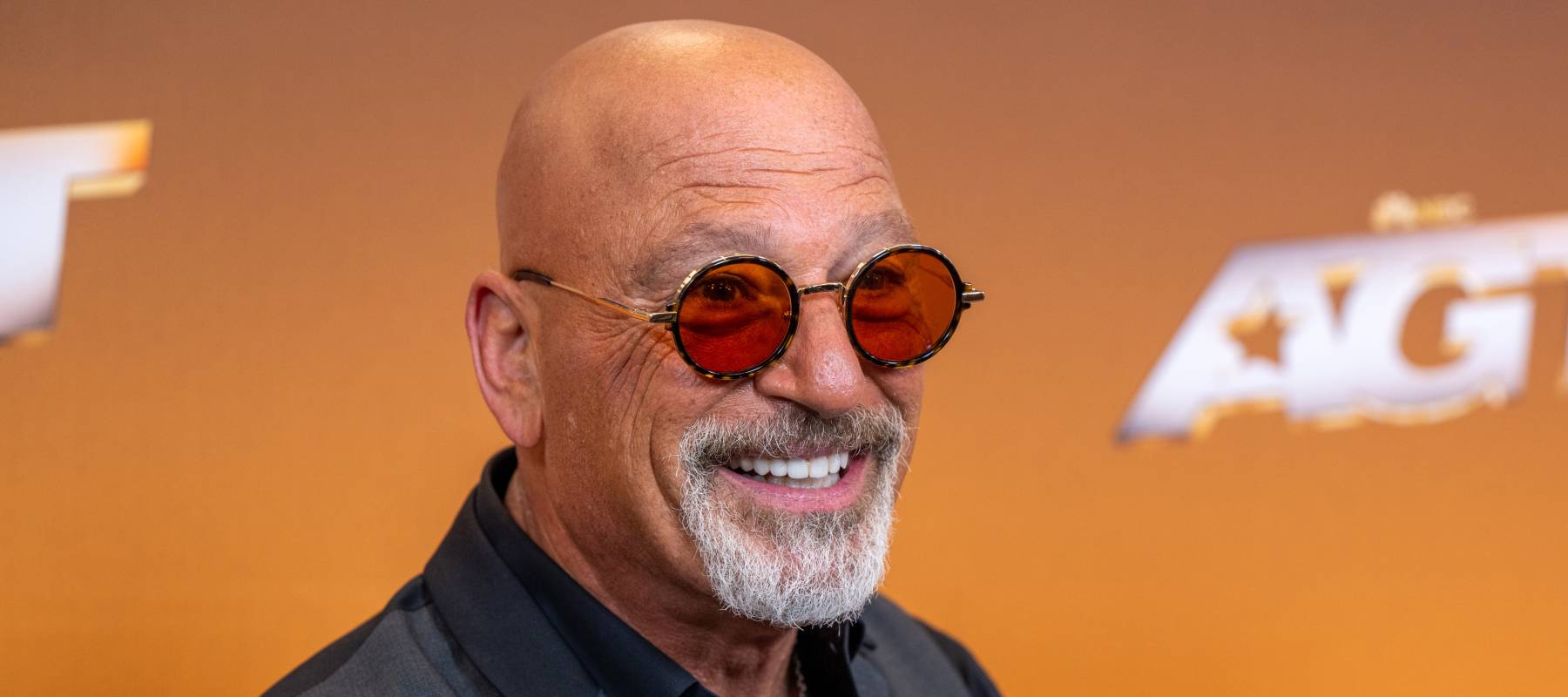 Howie Mandel, in his iconic red tinted glasses, grins broadly at the camera while dressed in a grey vest and black, open collared dress shirt.