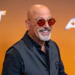 Howie Mandel, in his iconic red tinted glasses, grins broadly at the camera while dressed in a grey vest and black, open collared dress shirt.