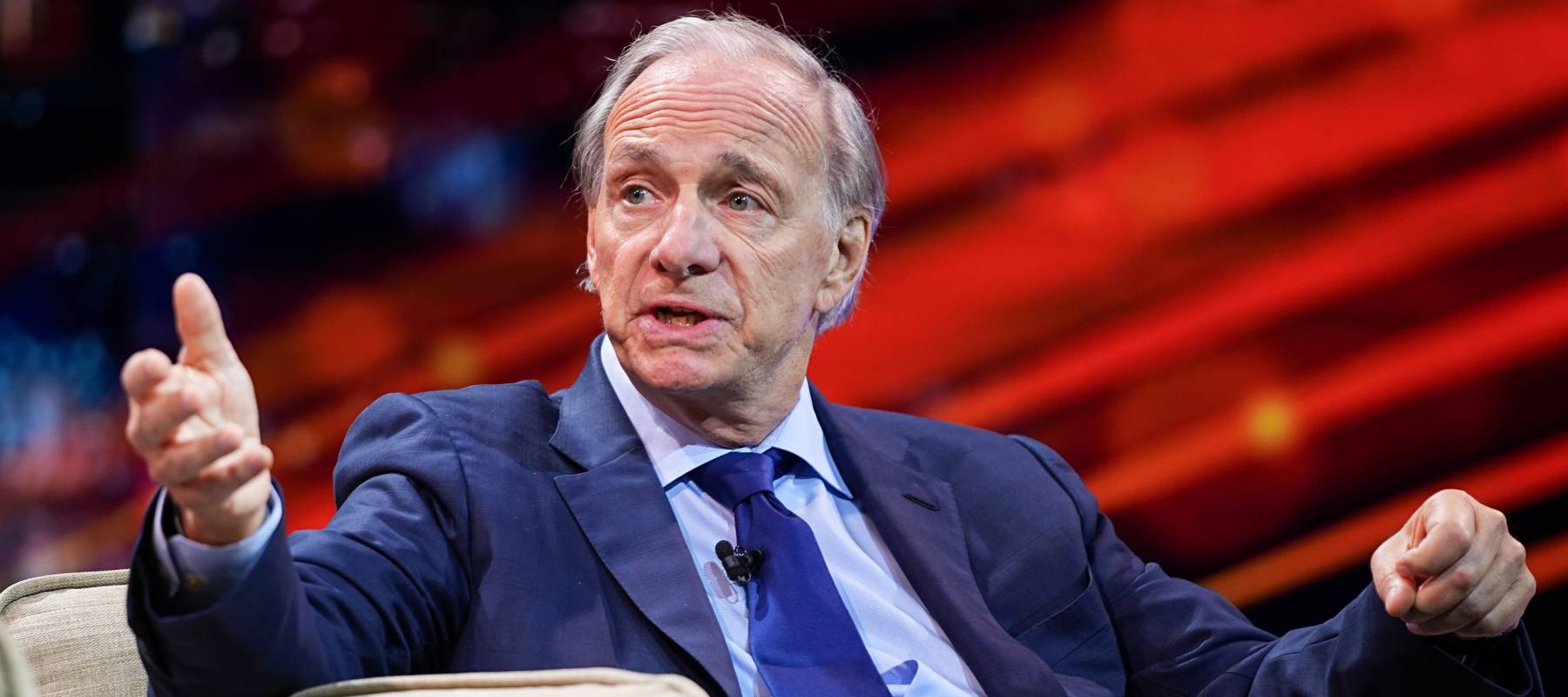 Ray Dalio, in a blue suit, shirt and tie, looks askew at the camera with one leg crossed over the other and with one hand gesturing outwards.