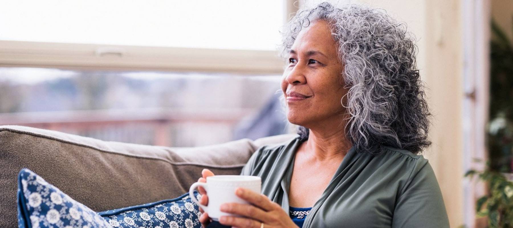 Senior woman holding coffee mug sitting on couch looking out window