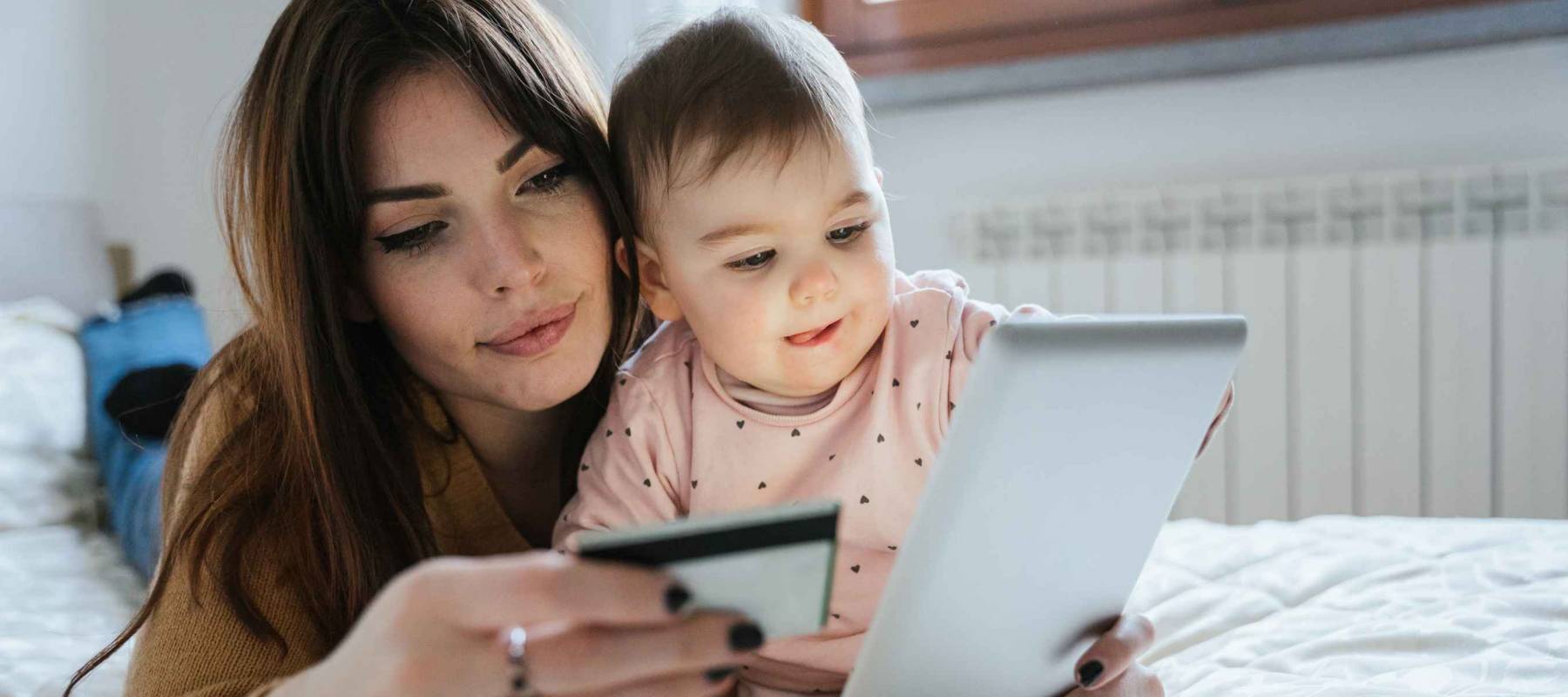 Portrait of a young mom with a baby girl lying on the bed  while buying products online with the tablet and holding her credit card in hand