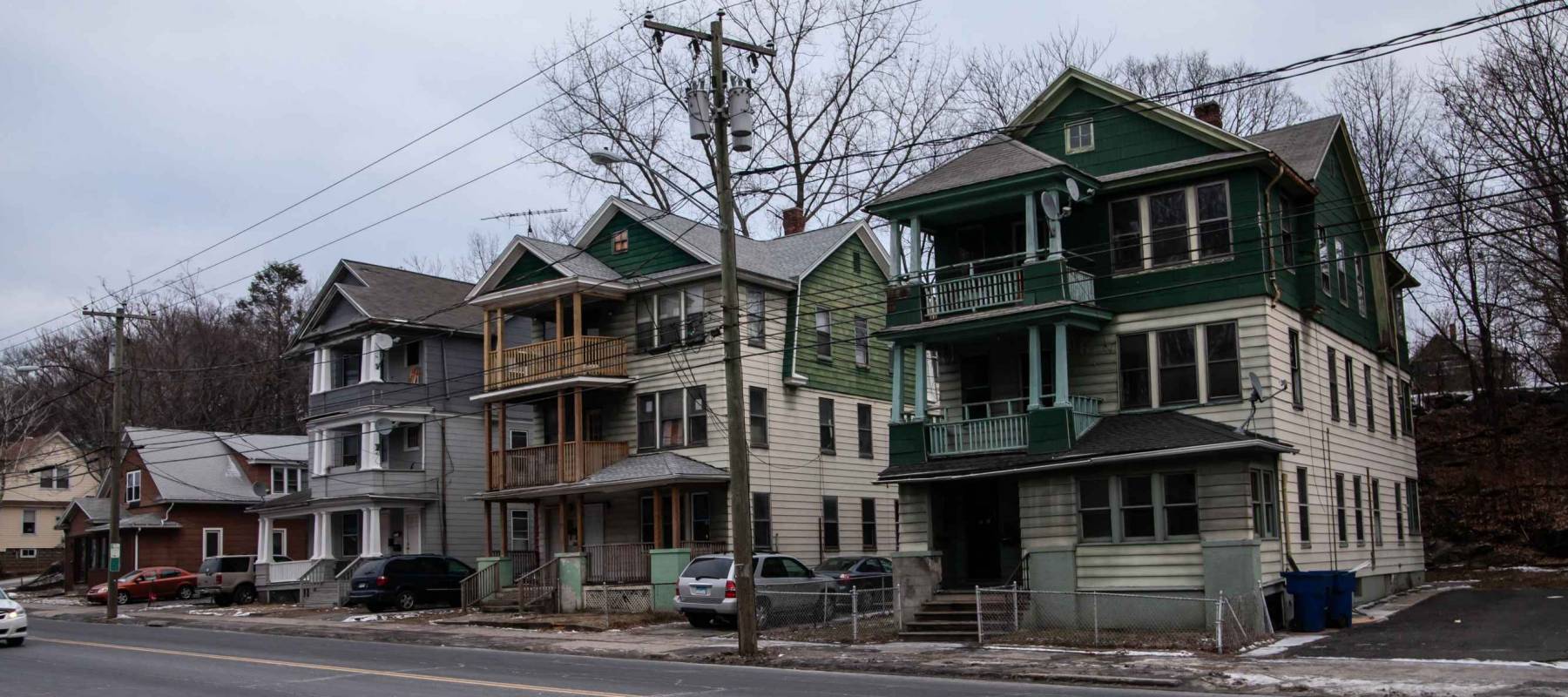 The run-down neighborhood with decrepit buildings surrounded by chain-link fences and old cars parked next to it on a gloomy, grey winter day.