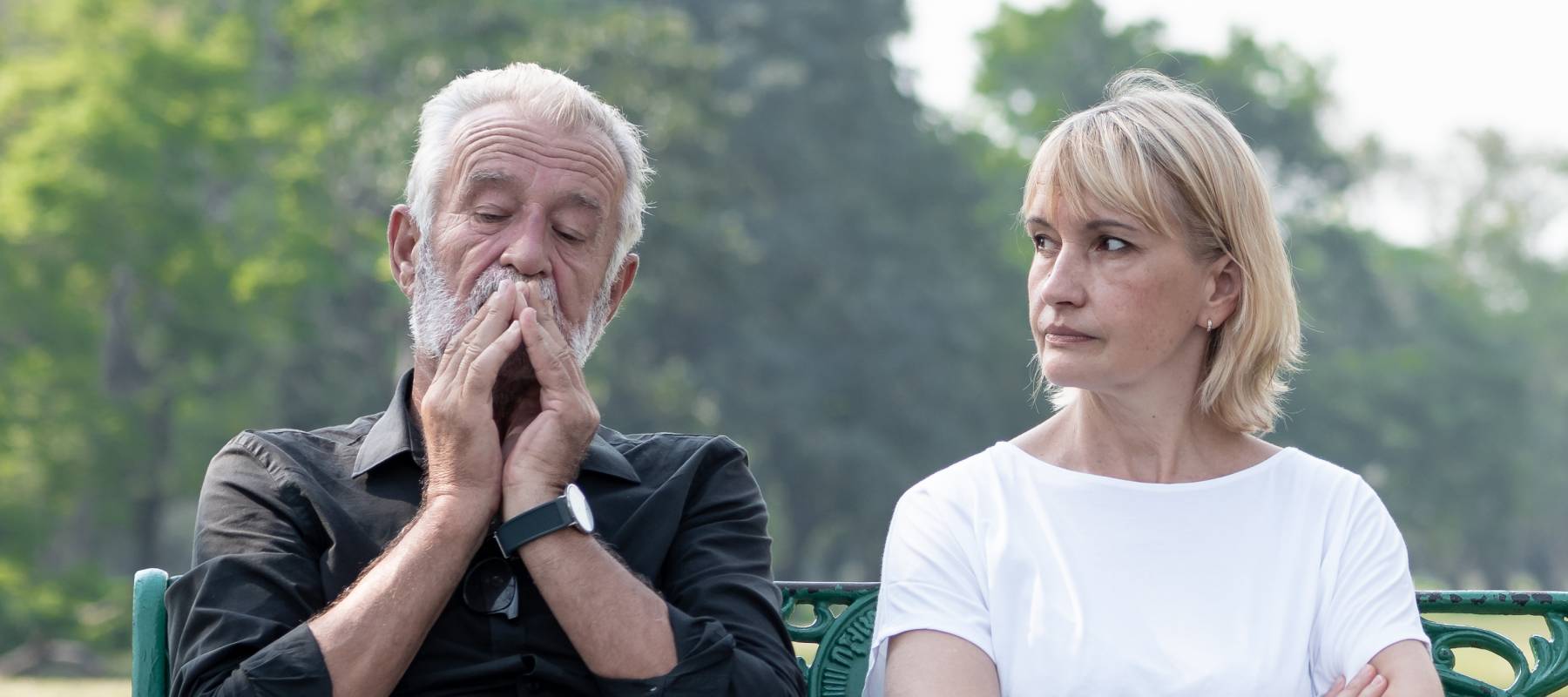 An older couple sit on a bench, looking concerned.