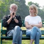 An older couple sit on a bench, looking concerned.