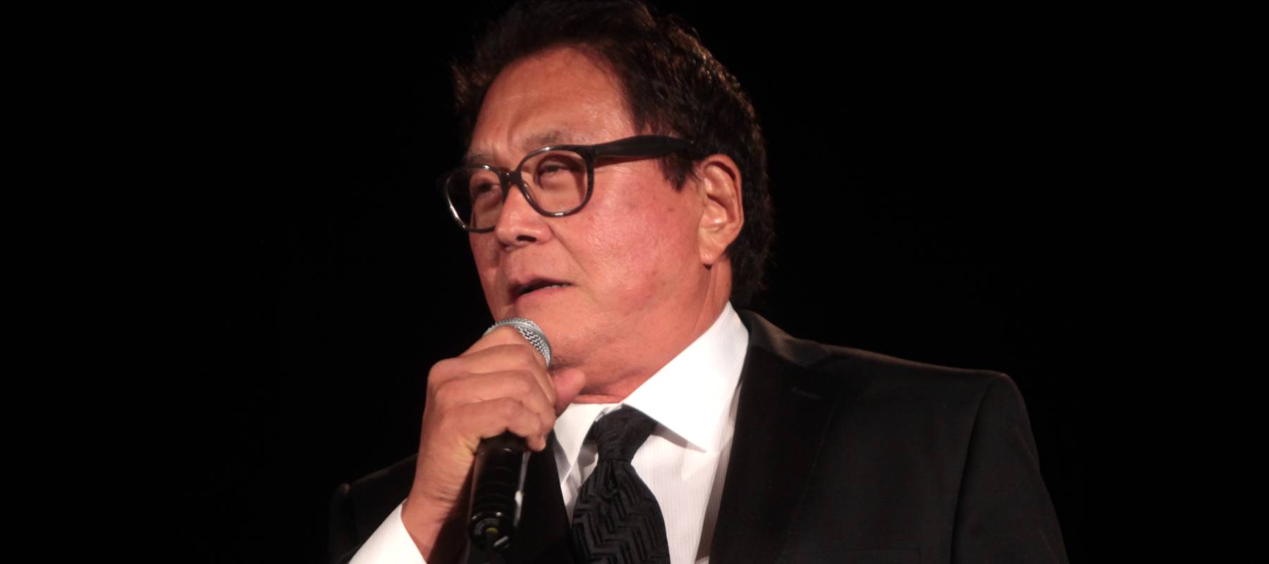 Robert Kiyosaki, in a pure white suit, looks at the audience at Freedomfest with wide eyes hidden behind clear glasses.