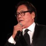 Robert Kiyosaki, in a pure white suit, looks at the audience at Freedomfest with wide eyes hidden behind clear glasses.