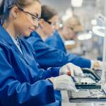Female Electronics Factory Worker in Blue Work Coat and Protective Glasses is Assembling Laptop's Motherboard with a Screwdriver. High Tech Factory Facility with Multiple Employees.