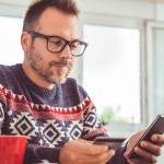 Men wearing blue sweater and eyeglasses holding credit card and using laptop at home office during christmas holidays