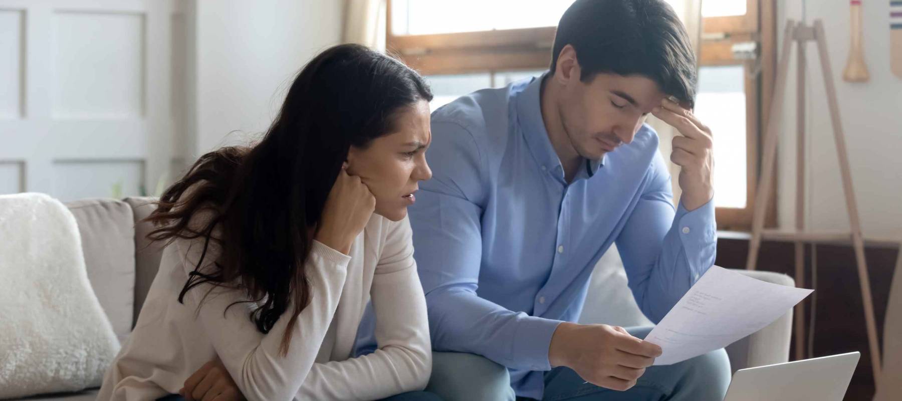 Man and woman sitting on a couch, looking distressed, holding papers