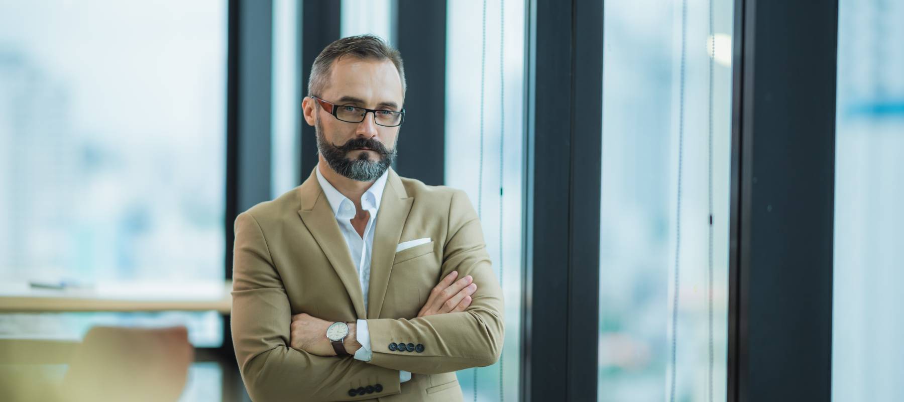 a bearded middle-aged Caucasian man with arms crossed standing in front of a window in a high-rise building