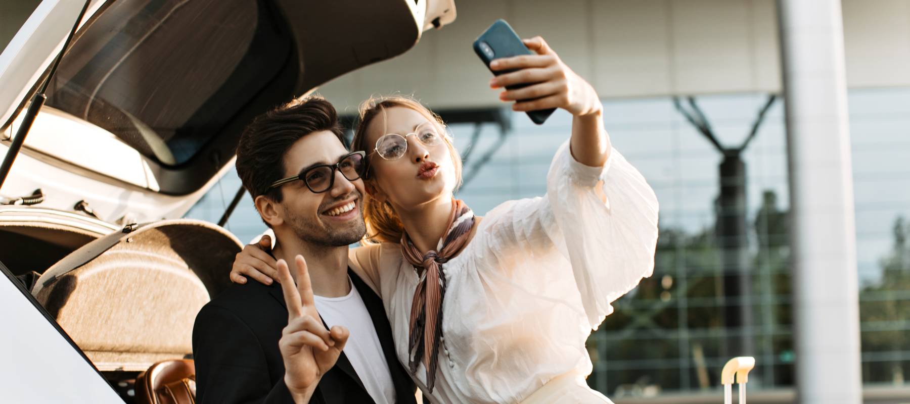 a young, wealthy Caucasian couple taking a selfie outside an airport