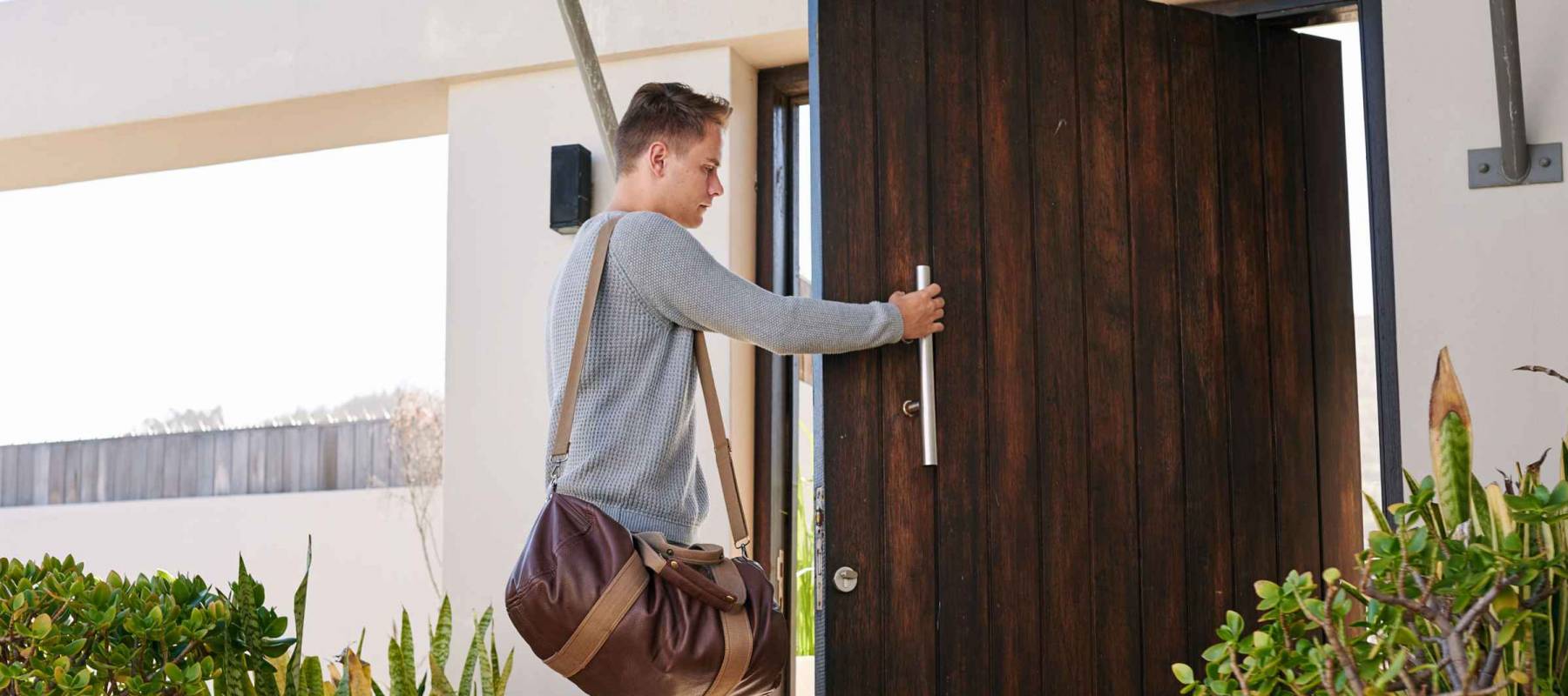 Young fashion designer busy closing his large wooden front door of his beautiful beach house with several plants planted along side the entrance