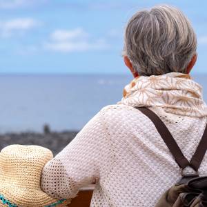 A senior woman with a backpack looks toward the sea's horizon.
