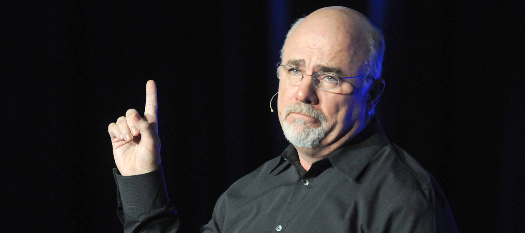 Dave Ramsey stands on stage in a black shirt with a single finger raised in the air.