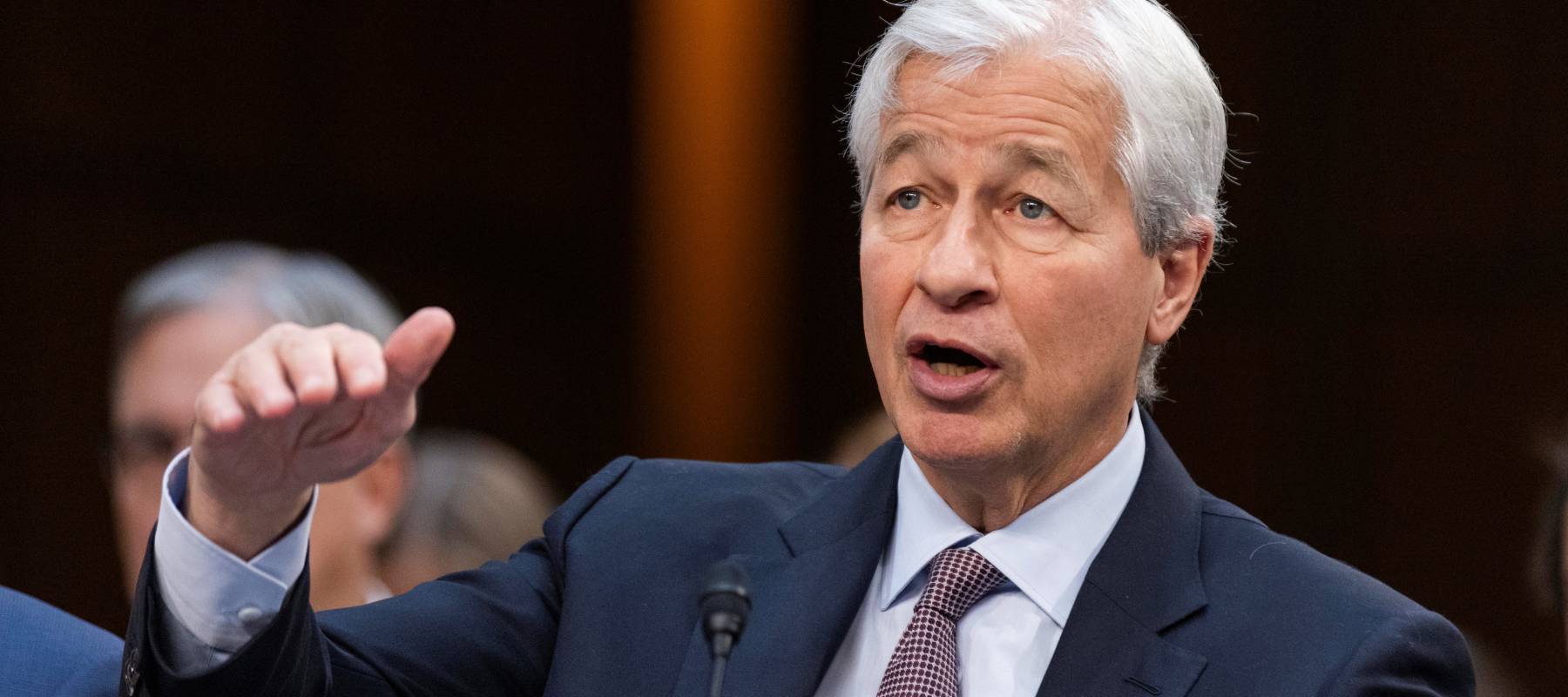 Jamie Dimon, in a blue suit jacket and pale blue dress shirt, speaks while gesturing broadly with his right hand.