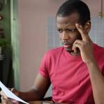 Young frustrated unhappy tired african american man with financial troubles, sitting at kitchen table with papers and calculator, reading documents