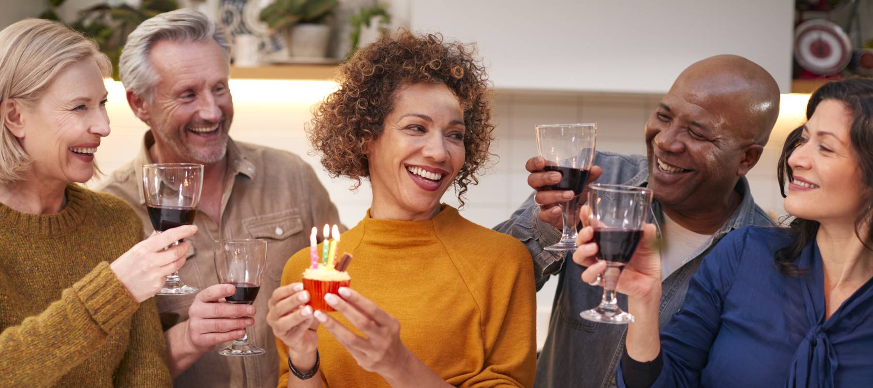 A group of friends clink glasses of wine as one holds a cupcake alight with three candles.
